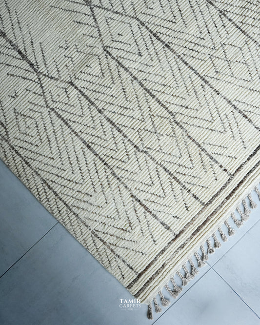 A close-up of a woven area rug with a cream and dark brown geometric herringbone pattern and fringed edge, laid on a gray tiled floor. A watermark for 'TAMIR CARPET' is visible at the bottom.