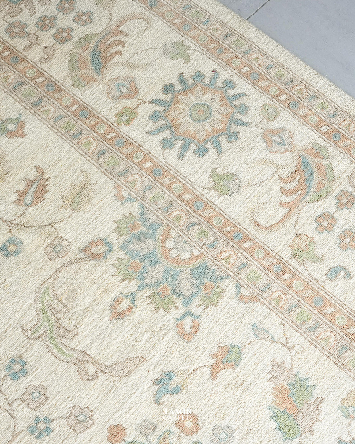 A close-up of a traditional area rug with an intricate floral and botanical pattern in muted beige, dusty blue, sage green, and soft orange tones on an off-white background.