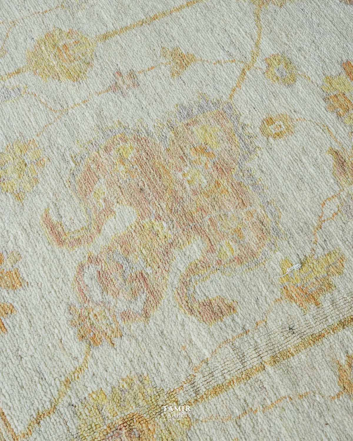 A close-up of a textured area rug featuring a faded traditional floral and scroll pattern in muted yellow, peach, and soft blue-gray tones on a warm off-white background.