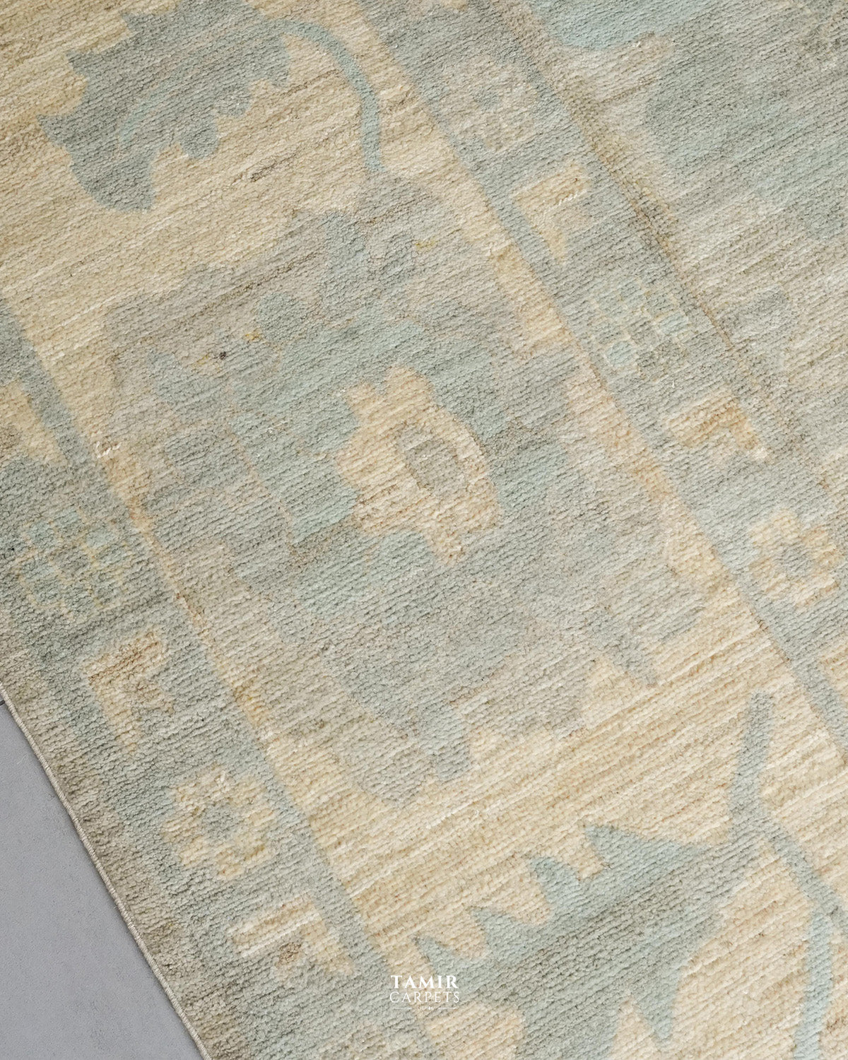 A close-up of a textured area rug with a distressed traditional pattern in muted sage green and warm beige, with a small TAMIR CARPETS logo at the bottom.
