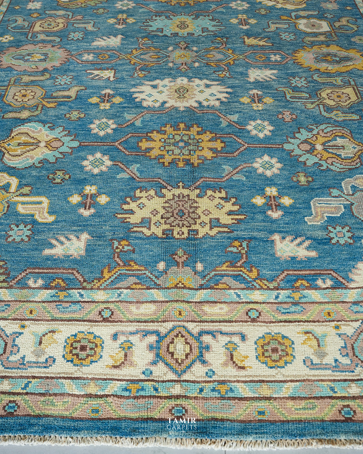 A close-up of a traditional patterned area rug with a turquoise blue field, cream borders, and motifs in beige, mustard yellow, and light blue. The rug features a woven texture and a small 'Tamir Carpets' logo watermark at the bottom center.