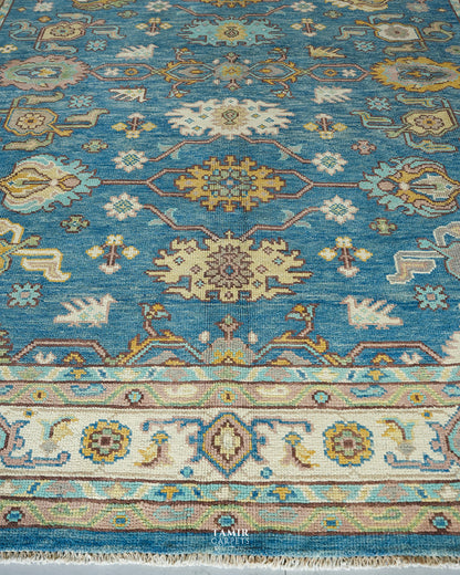 A close-up of a traditional patterned area rug with a turquoise blue field, cream borders, and motifs in beige, mustard yellow, and light blue. The rug features a woven texture and a small 'Tamir Carpets' logo watermark at the bottom center.