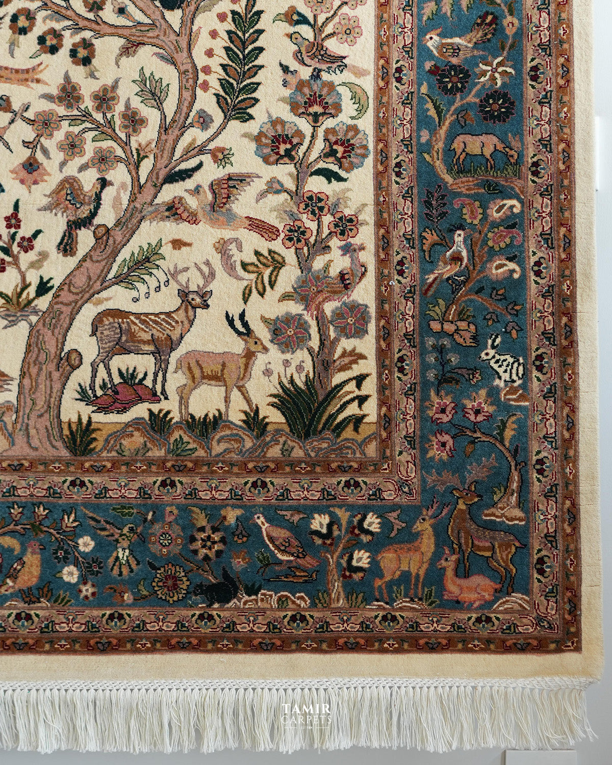 A close-up of a traditional oriental rug with a cream background, featuring a detailed forest scene with deer, birds, and floral motifs, framed by a blue and brown border with white fringe at the bottom.