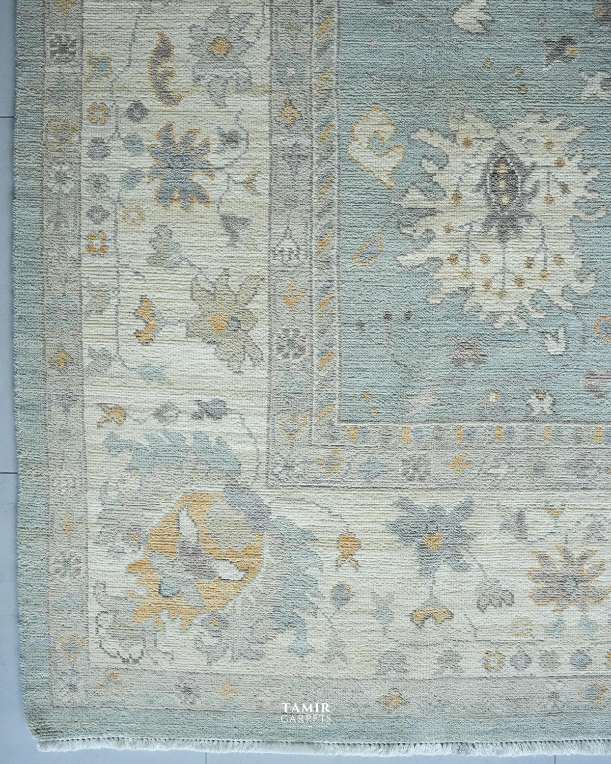A close-up of a traditional area rug with a dusty blue-green field, cream borders, and muted yellow and gray floral motifs. The rug features a fringed edge and a small 'TAMIR CARPETS' watermark at the bottom.