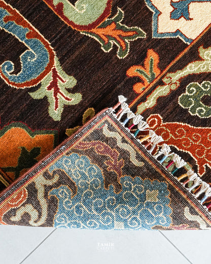A close-up of a patterned area rug with a dark brown base and traditional motifs in blue, orange, beige, and green. The corner of the rug is folded over to show the multicolored fringe.
