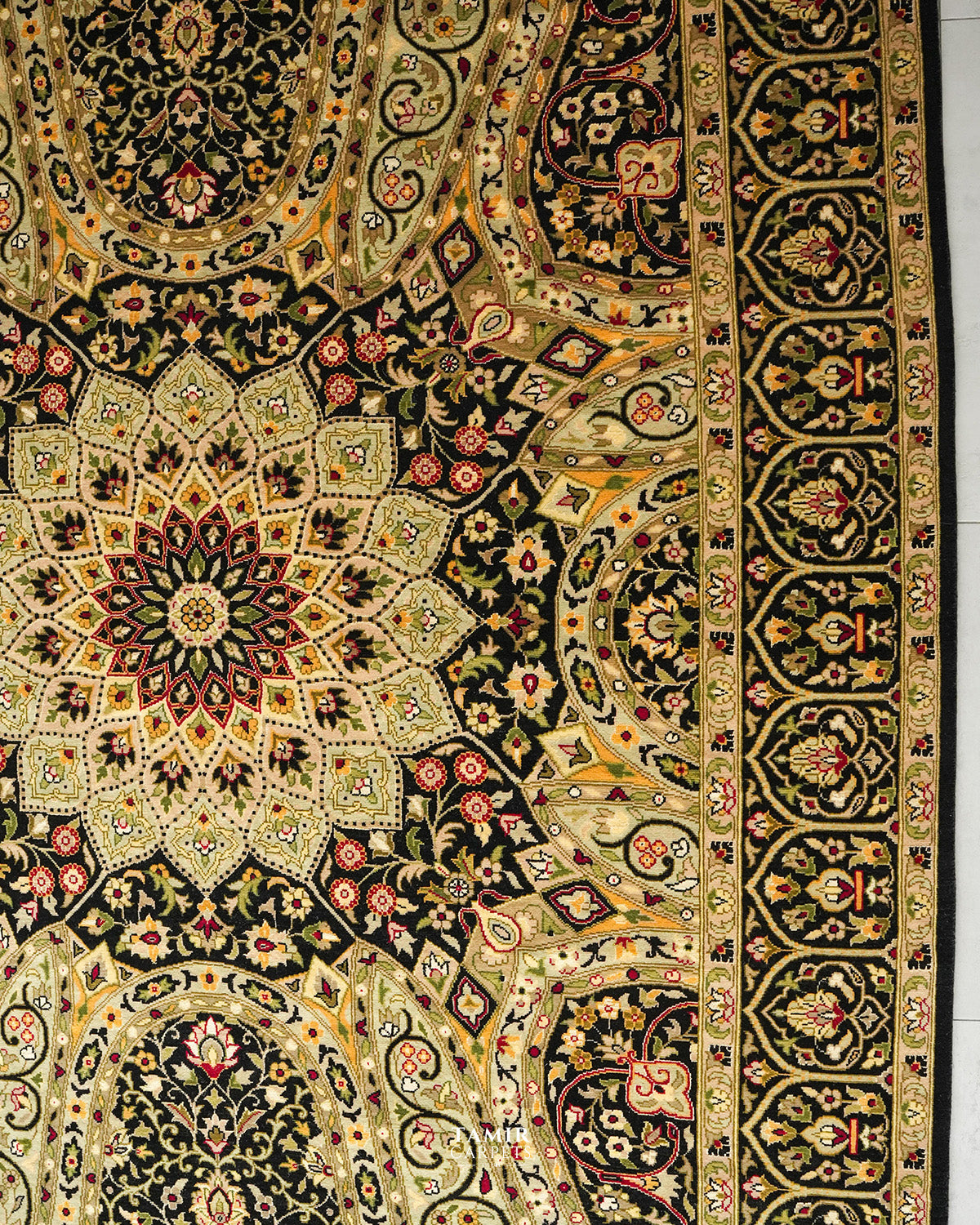 An ornate Persian-style area rug with a complex floral and geometric pattern in black, gold, beige, and red, shown laid flat with a white floor visible on the right edge.