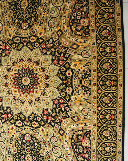 An ornate Persian-style area rug with a complex floral and geometric pattern in black, gold, beige, and red, shown laid flat with a white floor visible on the right edge.