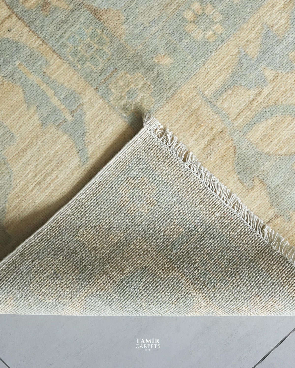 A close-up of a patterned area rug corner showing a light beige and pale blue-gray woven design with fringed edges, photographed on a neutral floor. The brand name TAMIR CARPETS is visible at the bottom.