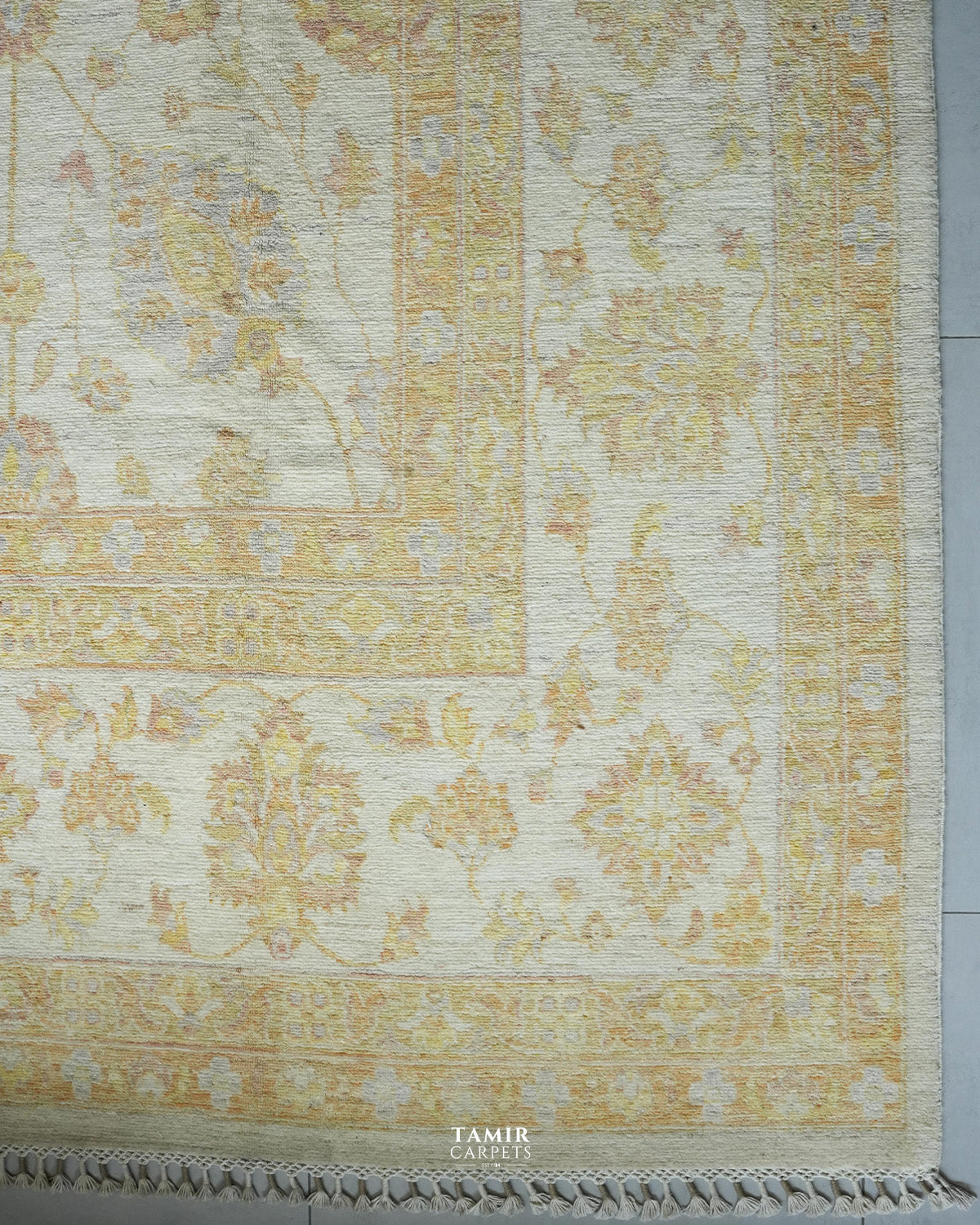 A close-up of a traditional area rug with an ornate floral pattern in soft beige, gold, and muted blue on an ivory background, featuring a fringed edge and a visible 'TAMIR CARPETS' watermark at the bottom.