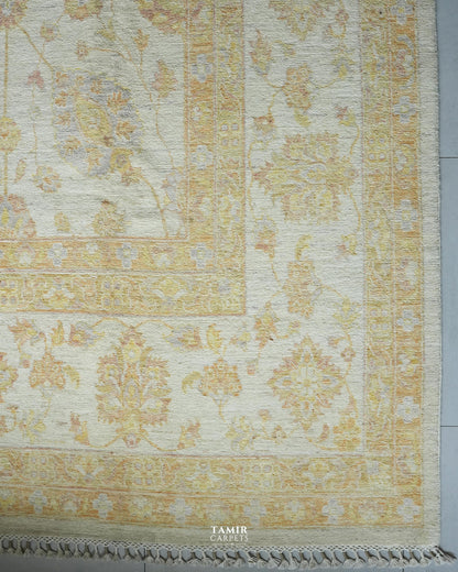 A close-up of a traditional area rug with an ornate floral pattern in soft beige, gold, and muted blue on an ivory background, featuring a fringed edge and a visible 'TAMIR CARPETS' watermark at the bottom.