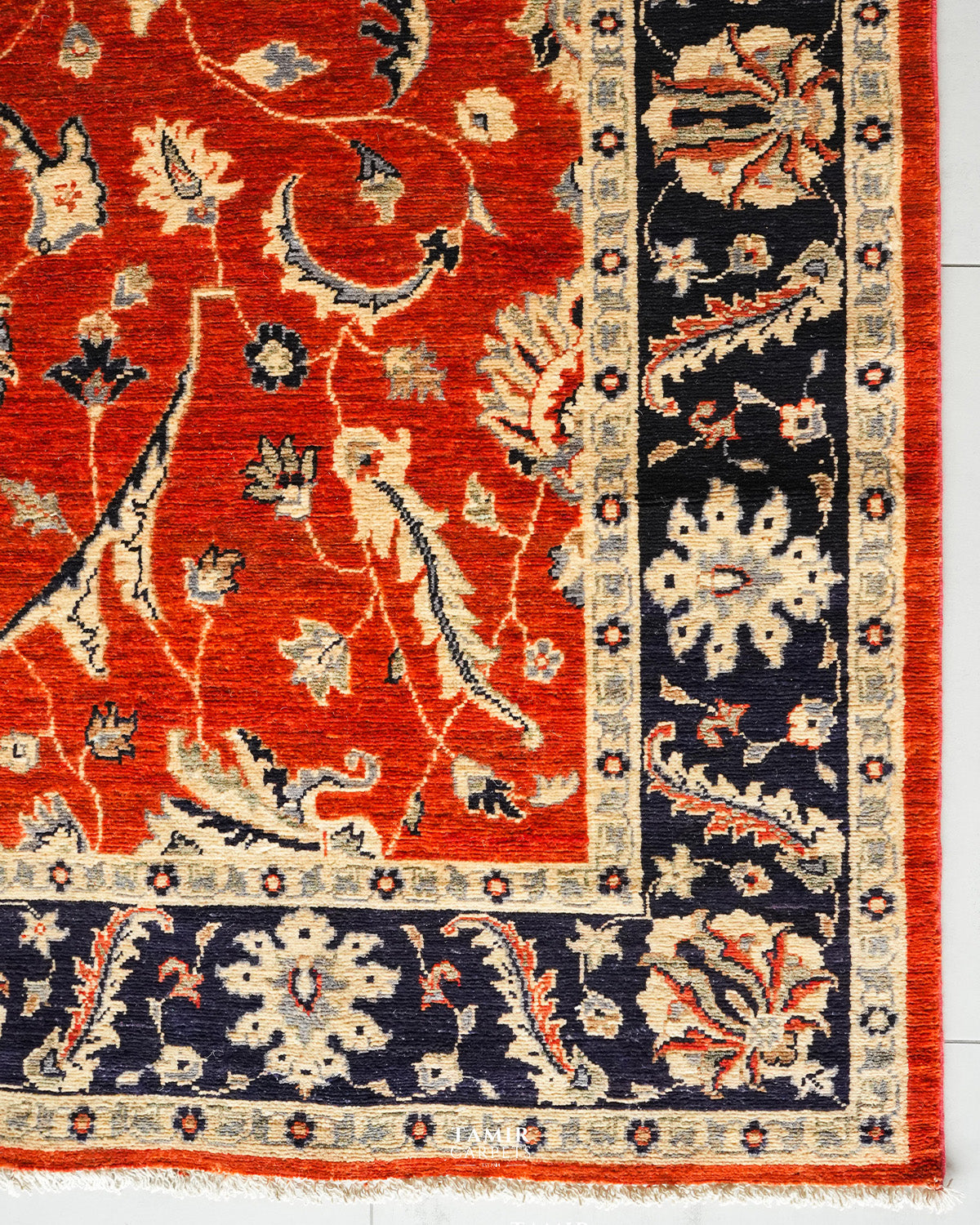 A close-up of a traditional patterned rug corner showing a red field with cream floral motifs, dark navy borders, and orange-red fringe.