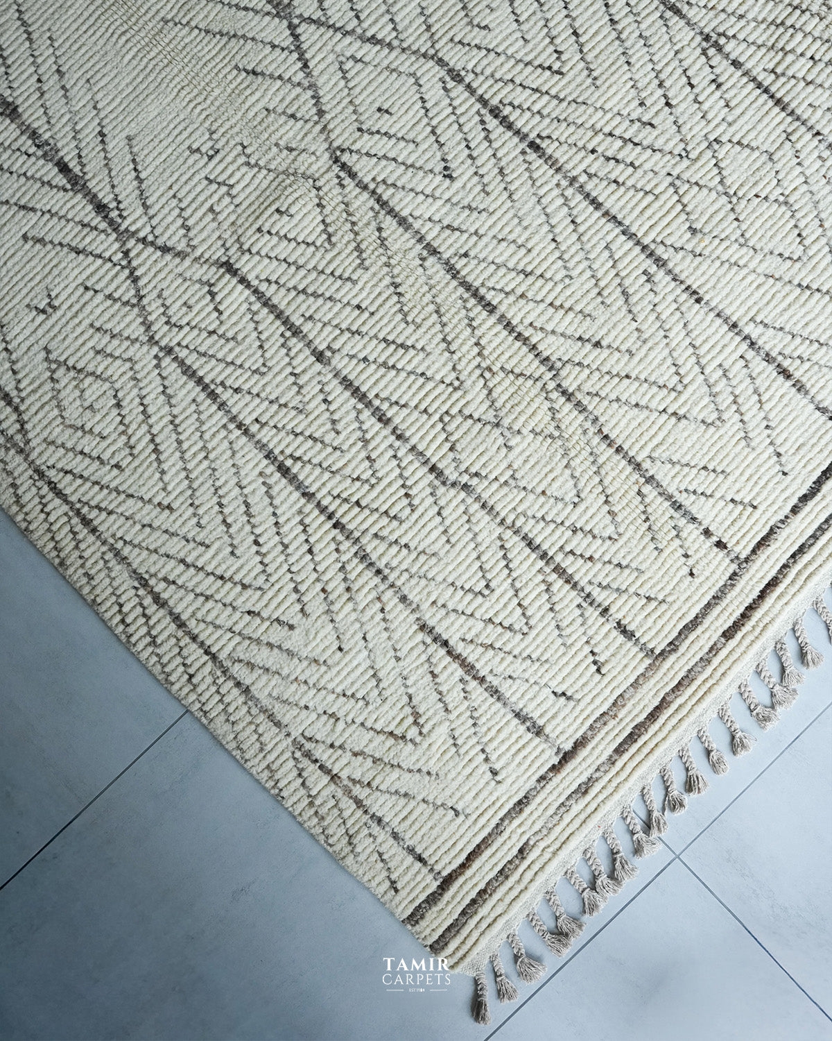 A close-up of a woven area rug with a cream and dark brown geometric herringbone pattern and fringed edge, laid on a gray tiled floor. A watermark for 'TAMIR CARPET' is visible at the bottom.