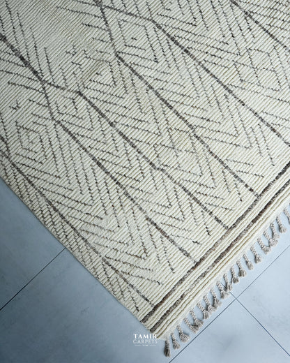 A close-up of a woven area rug with a cream and dark brown geometric herringbone pattern and fringed edge, laid on a gray tiled floor. A watermark for 'TAMIR CARPET' is visible at the bottom.
