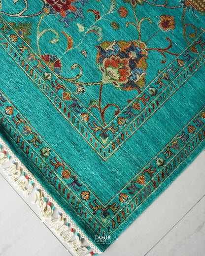 A close-up of a turquoise blue area rug with intricate floral and vine patterns in red, gold, and brown, featuring cream-colored fringe on the corner, laid out on a light gray floor.