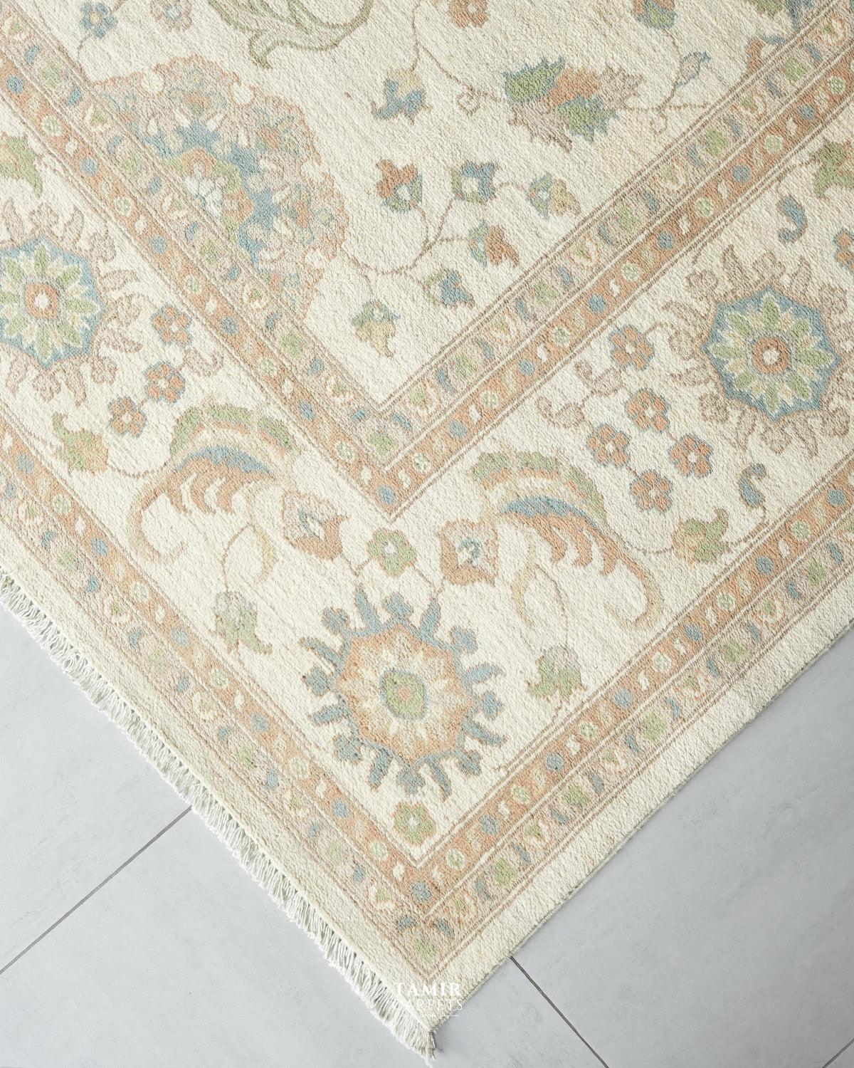A close-up corner view of a traditional area rug with an intricate floral and medallion pattern in beige, cream, muted blue, and soft green, laid on a light gray tiled floor.