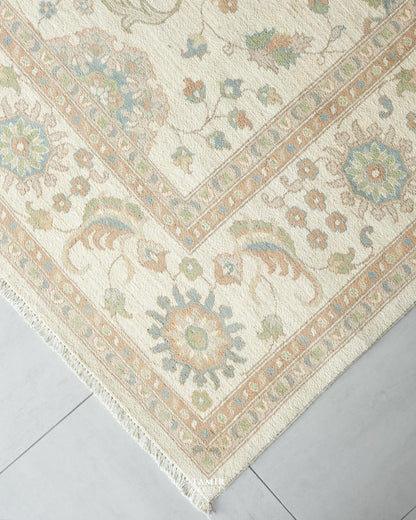 A close-up corner view of a traditional area rug with an intricate floral and medallion pattern in beige, cream, muted blue, and soft green, laid on a light gray tiled floor.