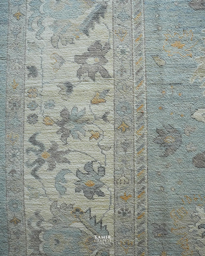 A close-up of a textured area rug featuring a traditional floral pattern in muted shades of dusty blue, gray, and beige with small gold accents.