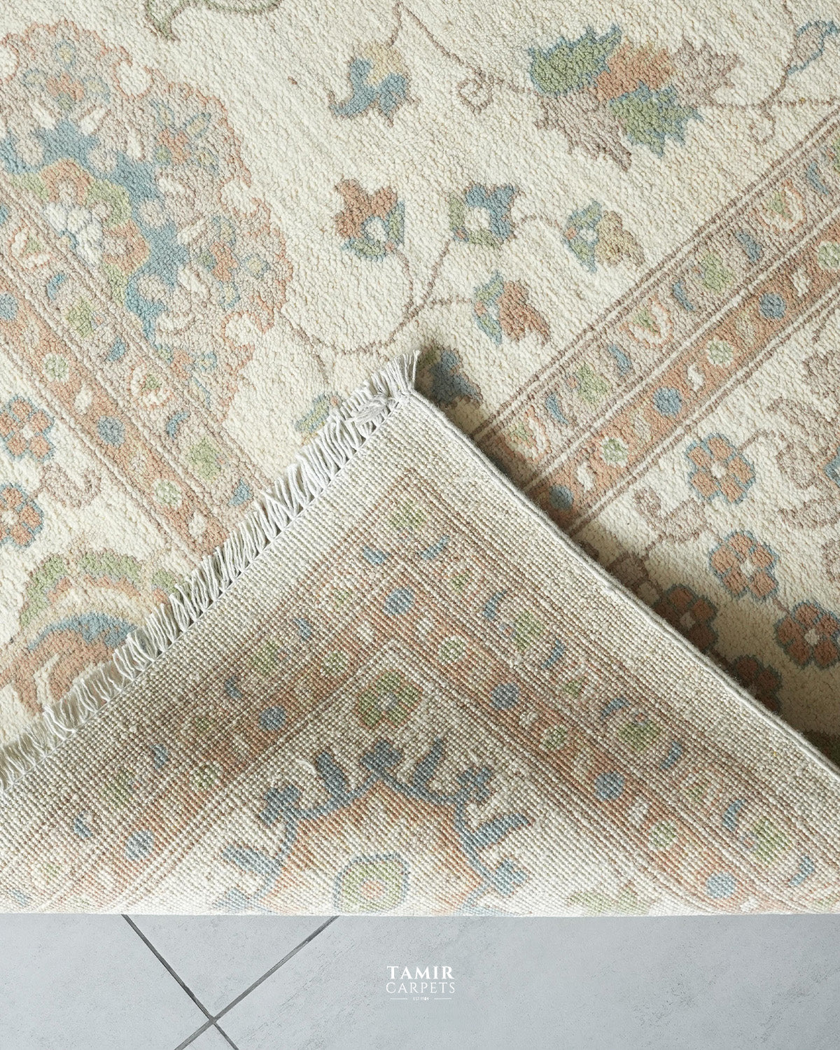 A close-up of a traditional patterned area rug with cream, beige, and muted blue-green tones, showing the corner and fringe detail. A tiled floor is visible at the bottom, and the watermark 'TAMIR CARPETS' appears at the bottom center.
