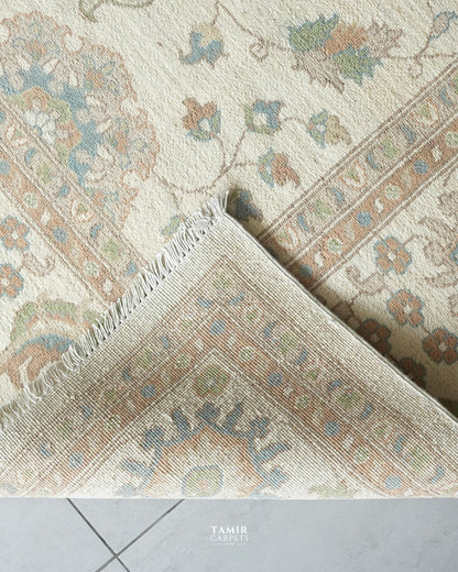 A close-up of a traditional patterned area rug with cream, beige, and muted blue-green tones, showing the corner and fringe detail. A tiled floor is visible at the bottom, and the watermark 'TAMIR CARPETS' appears at the bottom center.