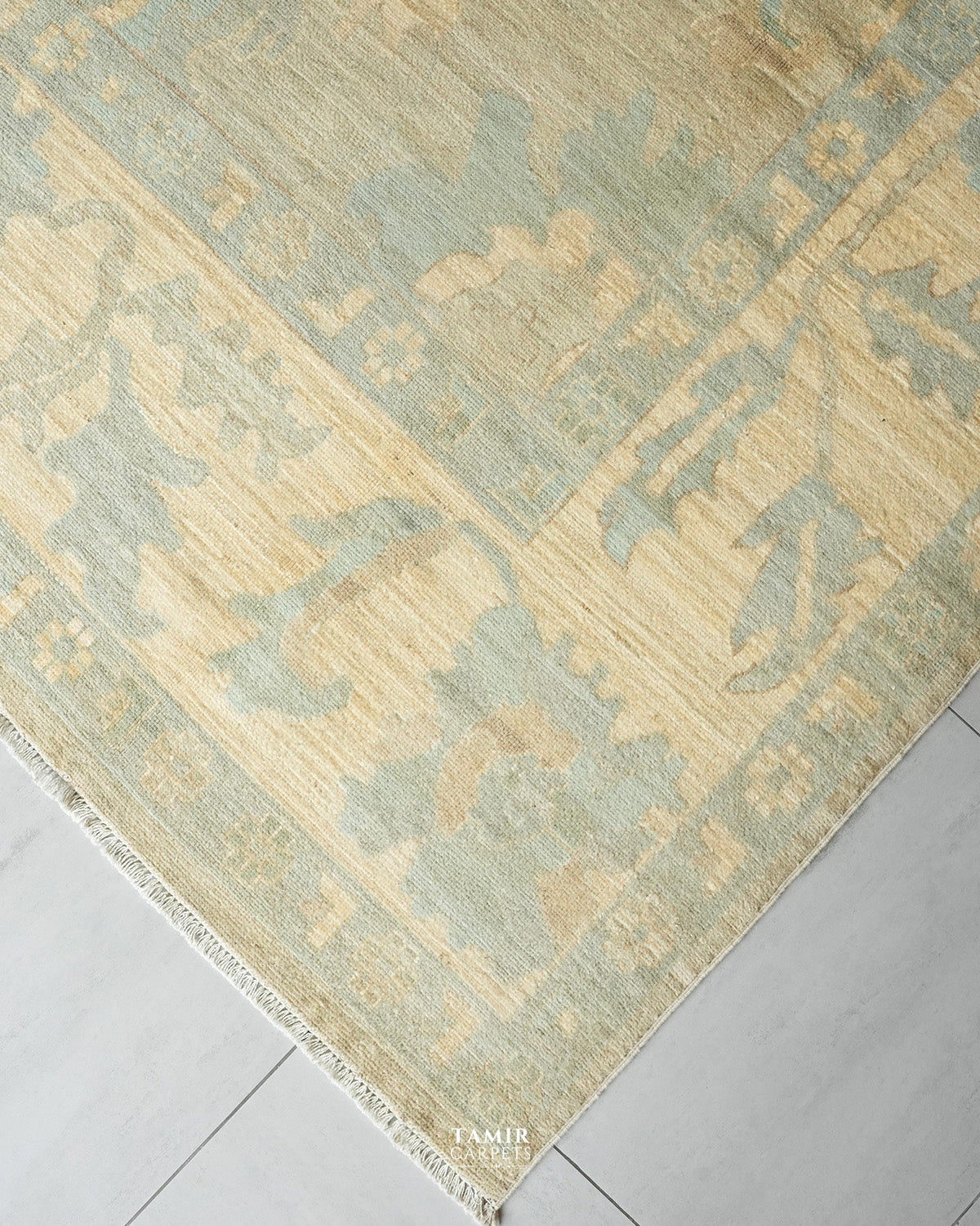 A close-up of a traditional area rug with a distressed, faded pattern in sage green and beige, laid out on a light gray tiled floor.