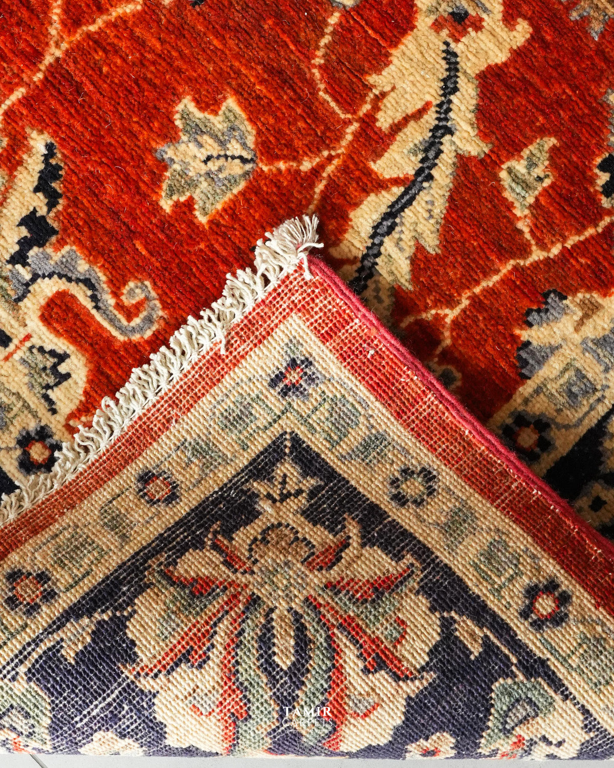 Close-up of a patterned area rug corner showing a red field with cream floral motifs and a navy blue corner panel with cream and red detailing, plus white fringe.