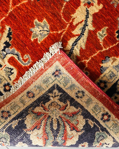 Close-up of a patterned area rug corner showing a red field with cream floral motifs and a navy blue corner panel with cream and red detailing, plus white fringe.