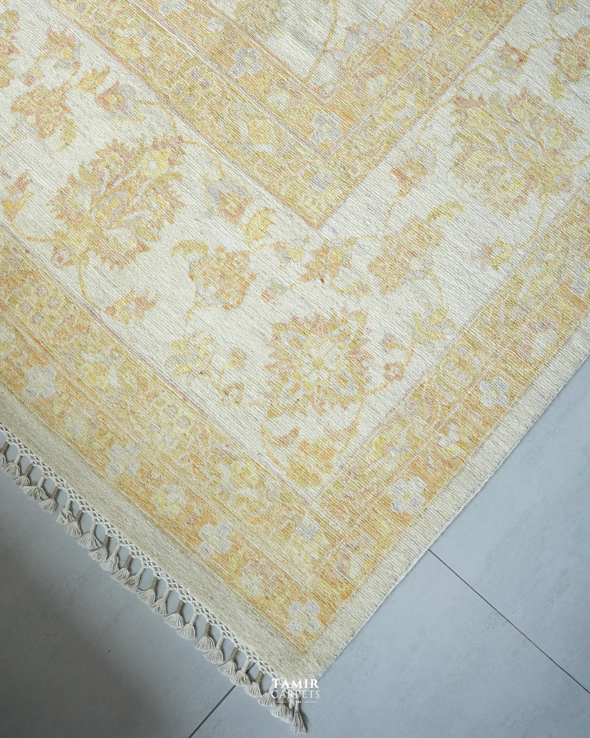 A close-up of a traditional area rug with an ornate floral pattern in pale yellow and cream, featuring a decorative fringed edge, laid out on a light gray tiled floor.