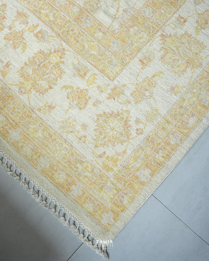 A close-up of a traditional area rug with an ornate floral pattern in pale yellow and cream, featuring a decorative fringed edge, laid out on a light gray tiled floor.