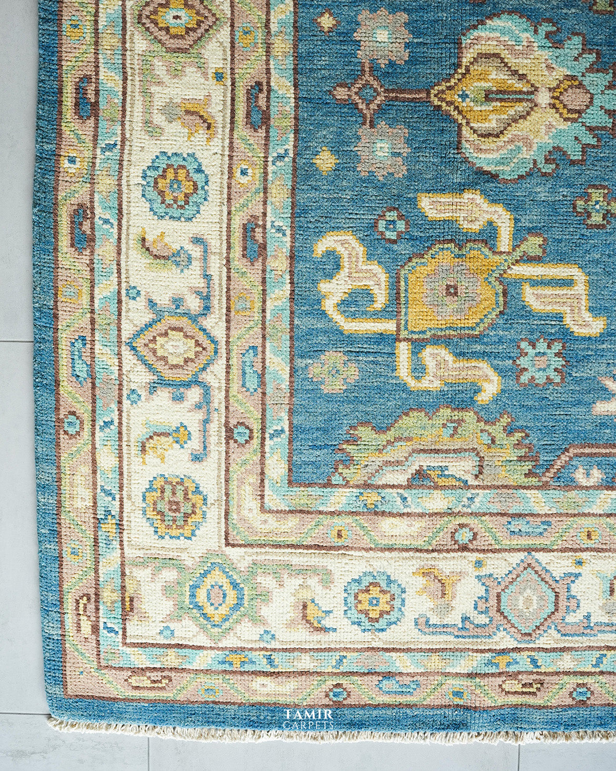 Close-up of a traditional patterned rug corner showing a blue field with cream, beige, and muted yellow motifs, surrounded by layered borders in cream and blue, with small white fringe at the bottom.
