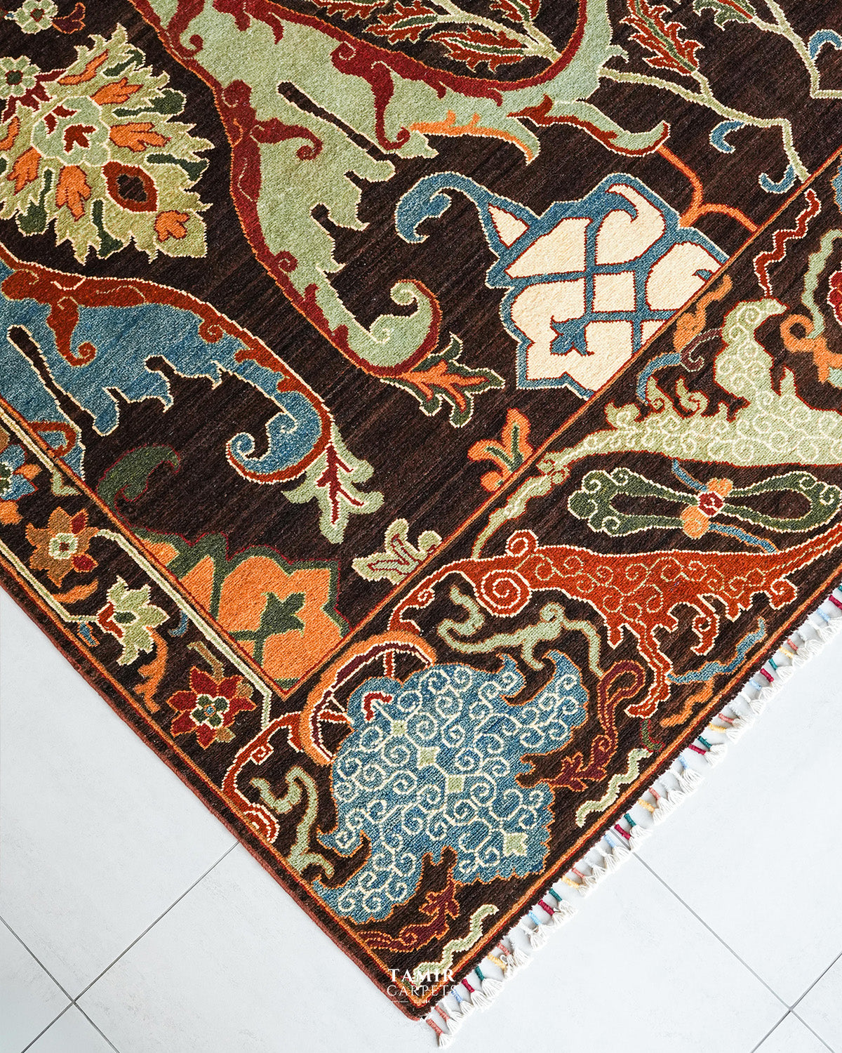 A close-up of an ornate patterned area rug corner, showing a dark brown field with cream, orange, light blue, and olive motifs, plus a decorative border and small fringe tassels, photographed on a light tiled floor.