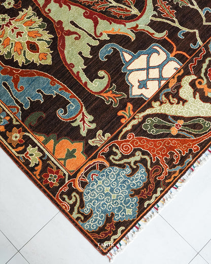 A close-up of an ornate patterned area rug corner, showing a dark brown field with cream, orange, light blue, and olive motifs, plus a decorative border and small fringe tassels, photographed on a light tiled floor.