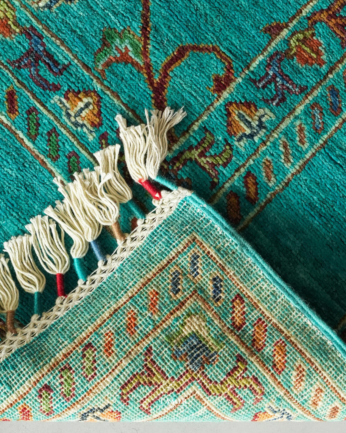 Close-up of a turquoise patterned rug with cream tassels and a decorative woven border, showing part of the folded corner and detailed textile texture.