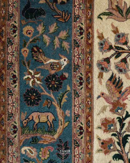 Close-up of a patterned area rug showing a blue central field with floral and bird motifs, a beige side panel with floral details, and ornate borders in rust, cream, and brown. A small TAMIR CARPETS logo appears near the bottom.
