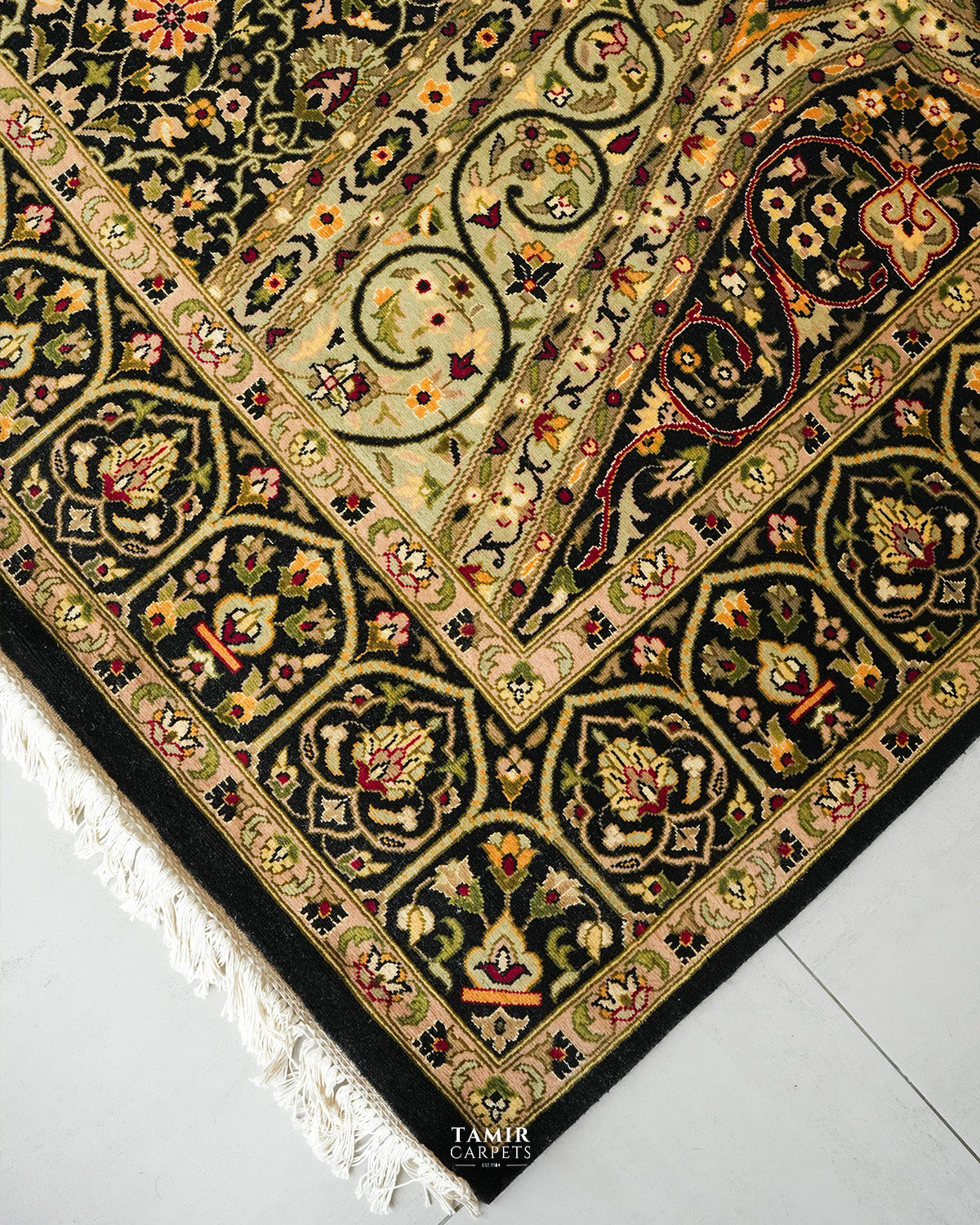 A close-up of an ornate patterned area rug corner, showing cream, black, green, and gold floral motifs with white fringe along the edge, photographed on a light floor. The brand name TAMIR CARPETS is visible at the bottom.
