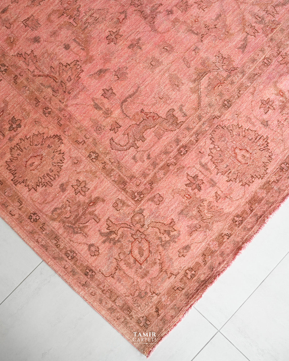 A dusty pink area rug with a faded traditional floral and medallion pattern, shown folded over on a light gray tiled floor. The brand name TAMIR CARPETS is visible in the bottom corner.