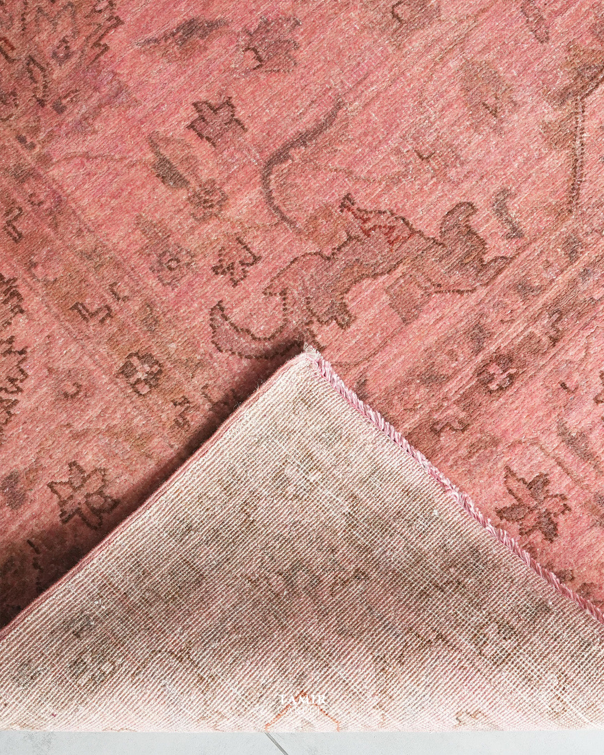 A close-up of a dusty pink area rug with a faded brown traditional motif, showing the corner folded over to reveal the woven texture and frayed edge.