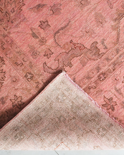 A close-up of a dusty pink area rug with a faded brown traditional motif, showing the corner folded over to reveal the woven texture and frayed edge.