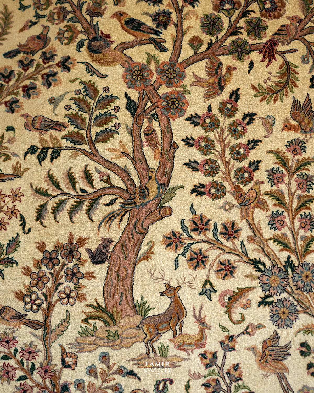 A close-up of a woven textile or rug pattern featuring a woodland scene with a tree trunk, birds, flowers, and a deer on a warm beige background. The word 'TAMIR CARPETS' appears near the bottom center.