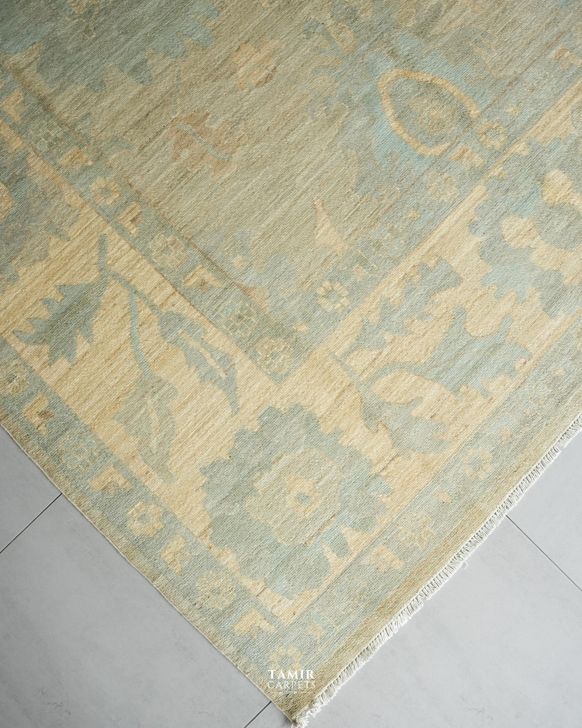 A close-up of a traditional area rug with a distressed medallion pattern in muted sage green and beige, shown on a light gray tiled floor. The brand name TAMIR CARPETS is visible in the bottom corner.