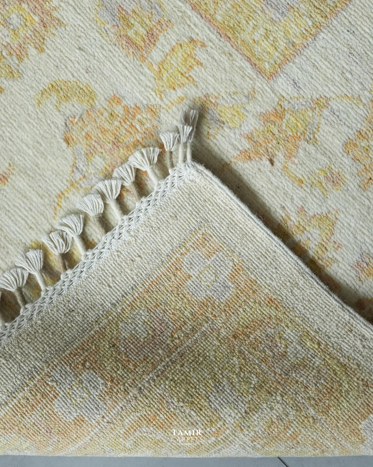 A close-up of a patterned area rug corner showing cream and beige tones with a subtle yellow floral motif and white tassel fringe along the edge.