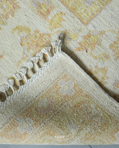 A close-up of a patterned area rug corner showing cream and beige tones with a subtle yellow floral motif and white tassel fringe along the edge.