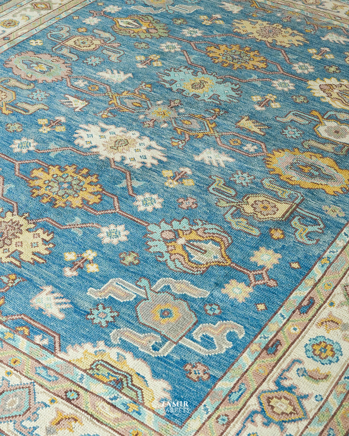 A close-up view of a traditional patterned area rug with a medium blue field, cream borders, and motifs in beige, yellow, and light blue.