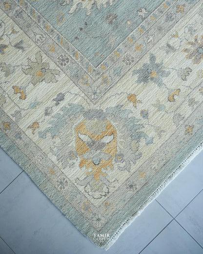 A close-up of a traditional area rug corner showing its detailed pattern and texture. The rug features a muted blue-gray field, cream borders, and accents of mustard yellow and gray in an ornate floral and medallion design.
