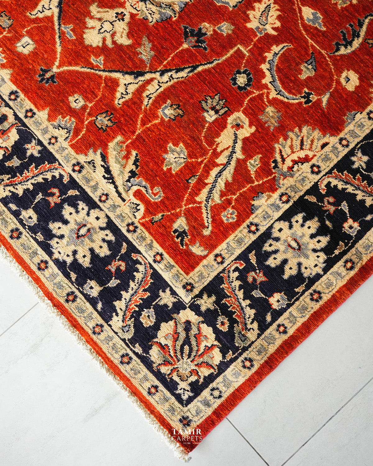 A close-up corner view of a traditional patterned area rug with a vibrant red field, dark navy blue border, and cream floral motifs, laid on a light gray marble floor.