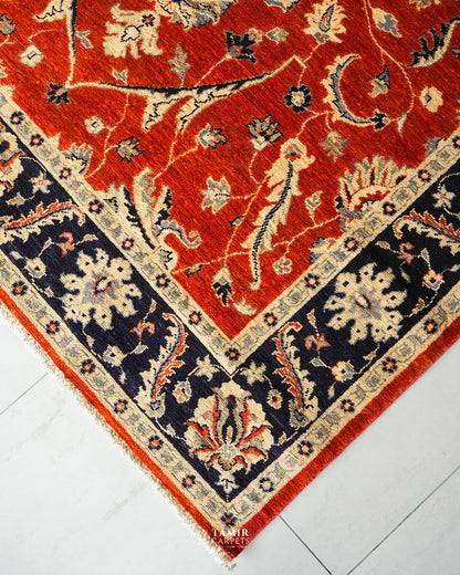 A close-up corner view of a traditional patterned area rug with a vibrant red field, dark navy blue border, and cream floral motifs, laid on a light gray marble floor.