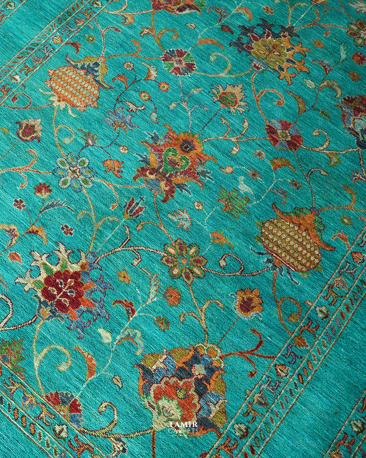 A close-up of a turquoise blue patterned rug with intricate floral motifs and ornate borders in red, gold, beige, and brown.