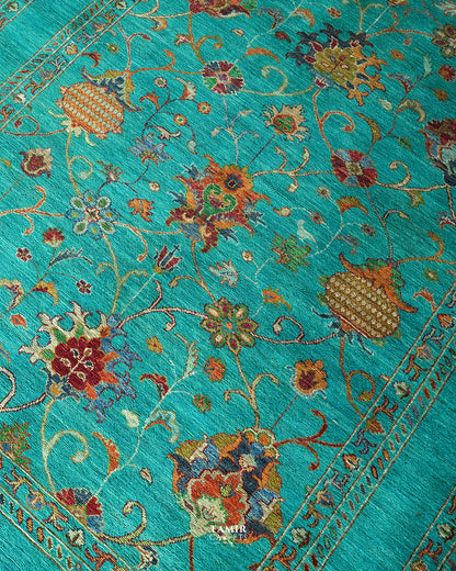 A close-up of a turquoise blue patterned rug with intricate floral motifs and ornate borders in red, gold, beige, and brown.