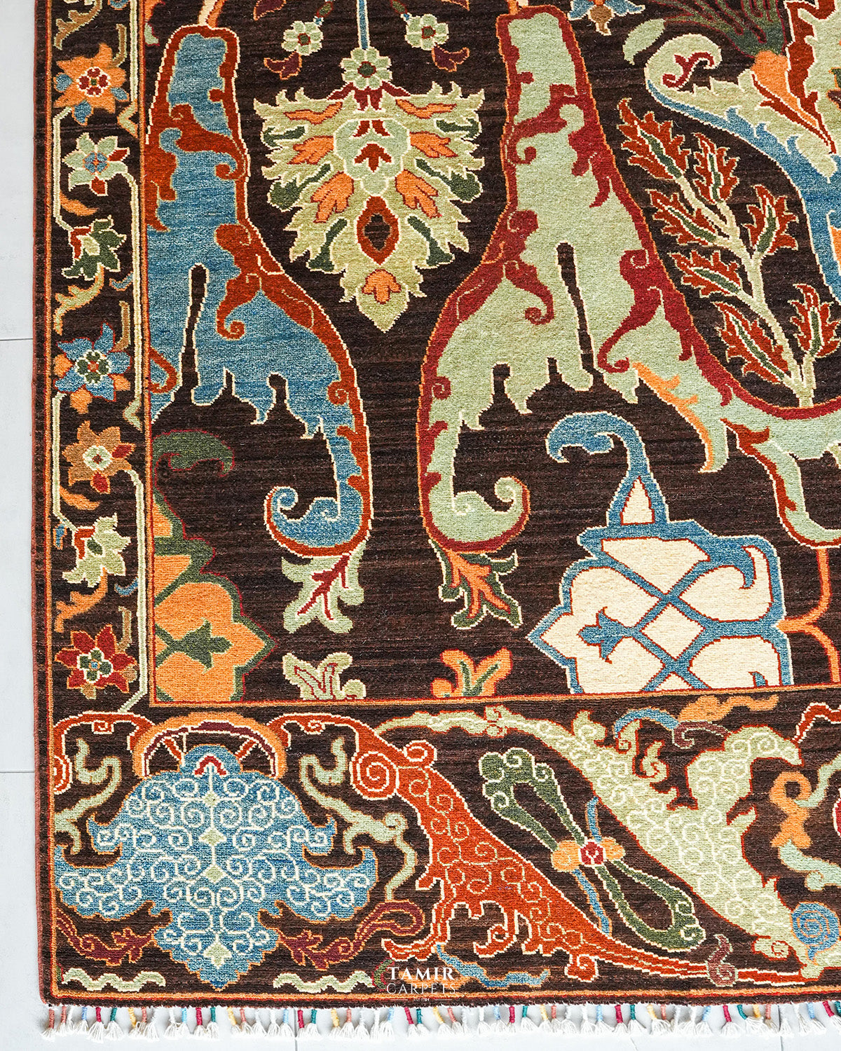 A close-up of an ornate patterned rug with a dark brown field and traditional motifs in blue, orange, beige, and cream, including stylized floral and scroll designs.