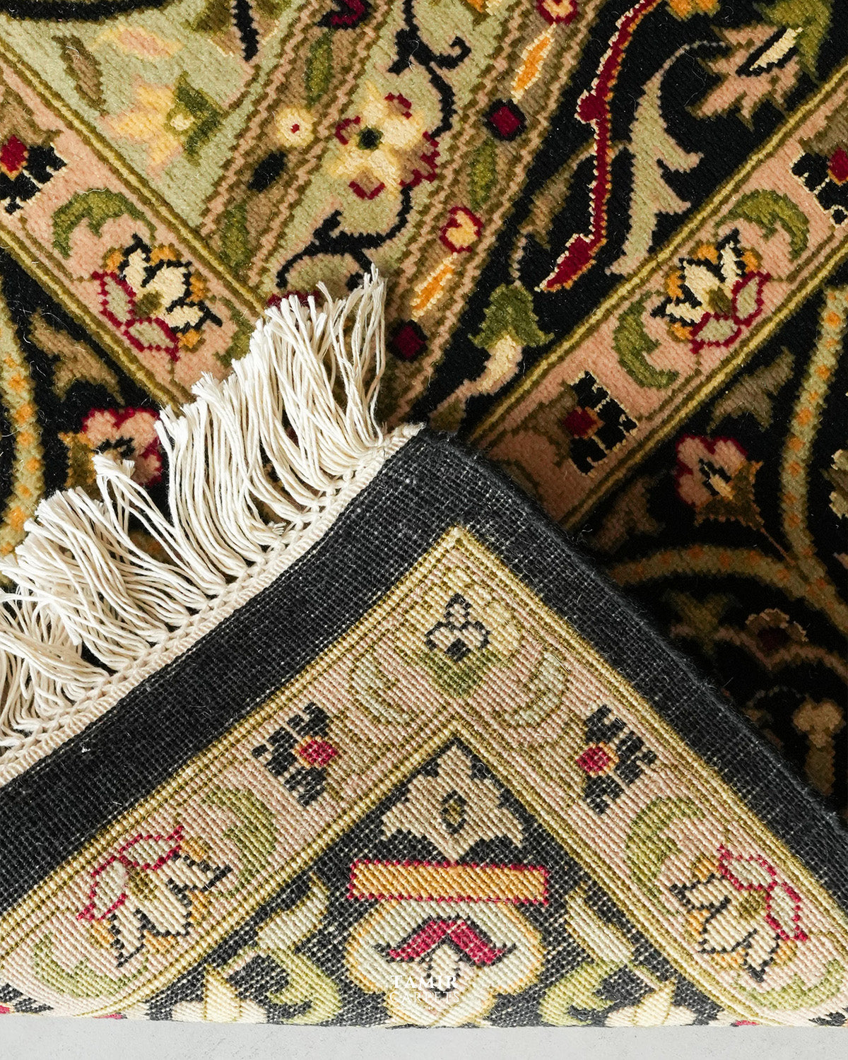 Close-up of an ornate patterned rug corner with cream fringe, featuring beige, black, green, red, and yellow woven floral and geometric motifs.