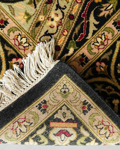 Close-up of an ornate patterned rug corner with cream fringe, featuring beige, black, green, red, and yellow woven floral and geometric motifs.